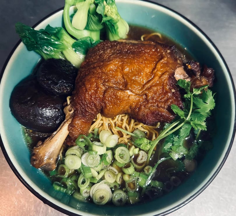 A bowl of duck noodle soup with bok choy, mushrooms, scallions, and cilantro.
