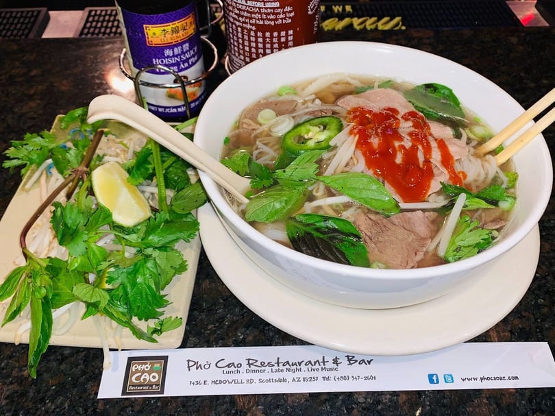 A bowl of Pho Cao pho with beef, noodles, and fresh herbs, alongside hoisin and sriracha.