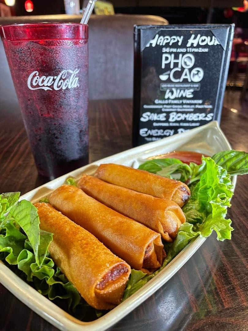 Four crispy spring rolls on lettuce with a Coca-Cola and a happy hour menu in the background.